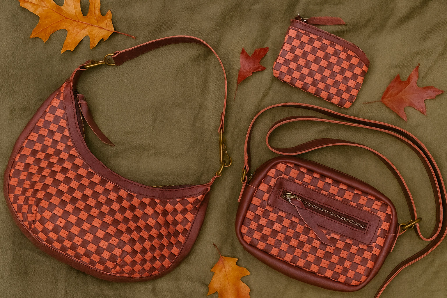 ShopYego Garnet Desert Rose collection flat lay featuring the Bella Crescent Bag, Gode Crossbody, and Shashemene Wallet. Handwoven leather in plum and dusty rose displayed on an olive green picnic blanket with fall leaves, artisan made in Ethiopia.
