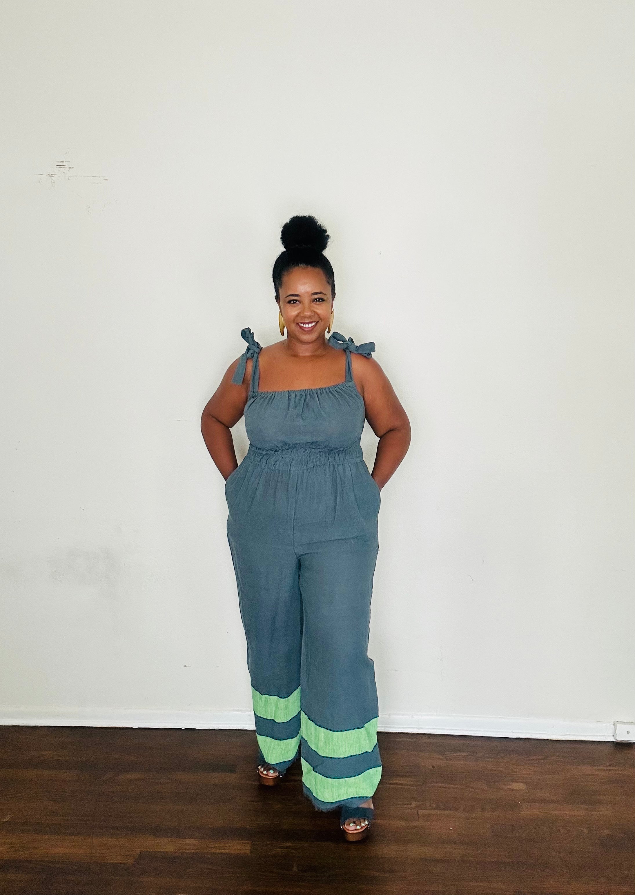 Handmade Blue Slate Sebie Jumpsuit in 100% cotton, featuring a flattering waist, adjustable straps, and deep pockets. Finished with a signature brown hem detailed with lime green accents for a bold, free-spirited LA style